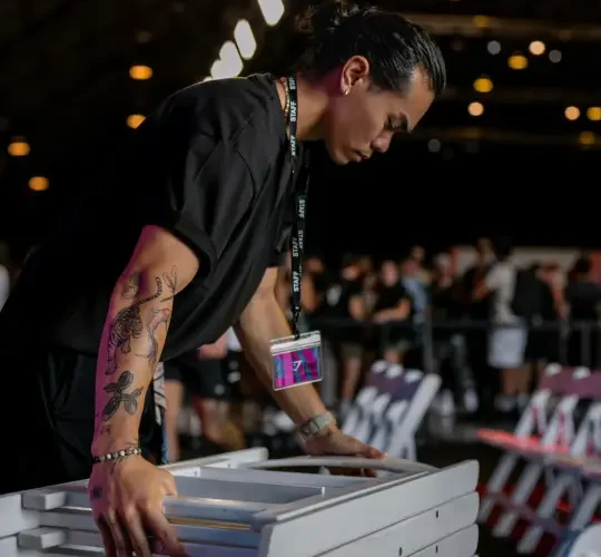 Premier Staff team member with a tattoo on their arm, leaning over a stack of white chairs at an event, wearing a black shirt and a lanyard with a badge.