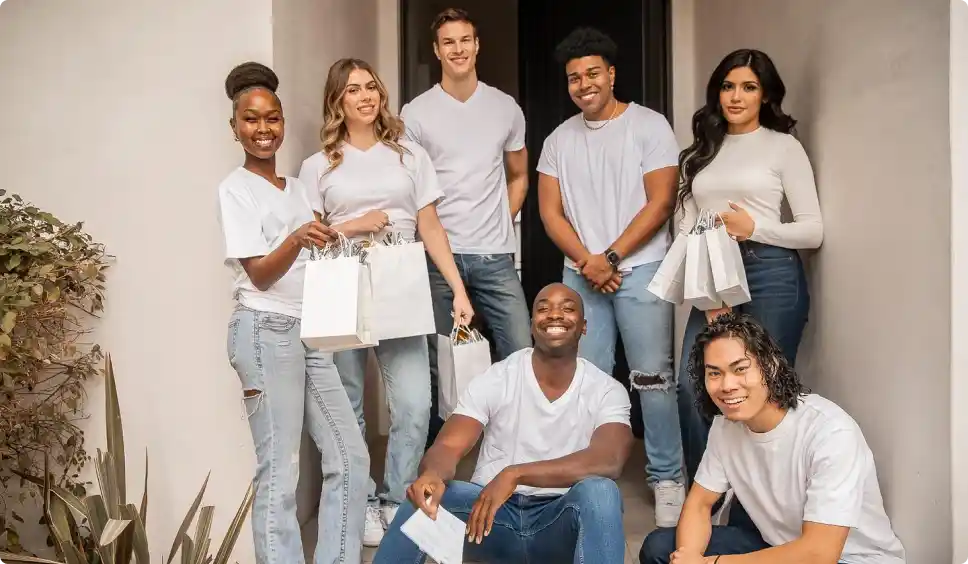 Group of seven Premier Staff members dressed in casual white tops and blue jeans, standing and sitting on a staircase outside, some holding white gift bags.