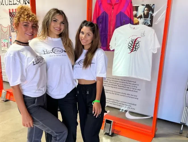 Premier Staff brand ambassadors wearing white t-shirts with 'Her Rise' and 'She is never done' printed on them, posing together at an indoor event with a display case in the background showcasing a t-shirt and jacket.