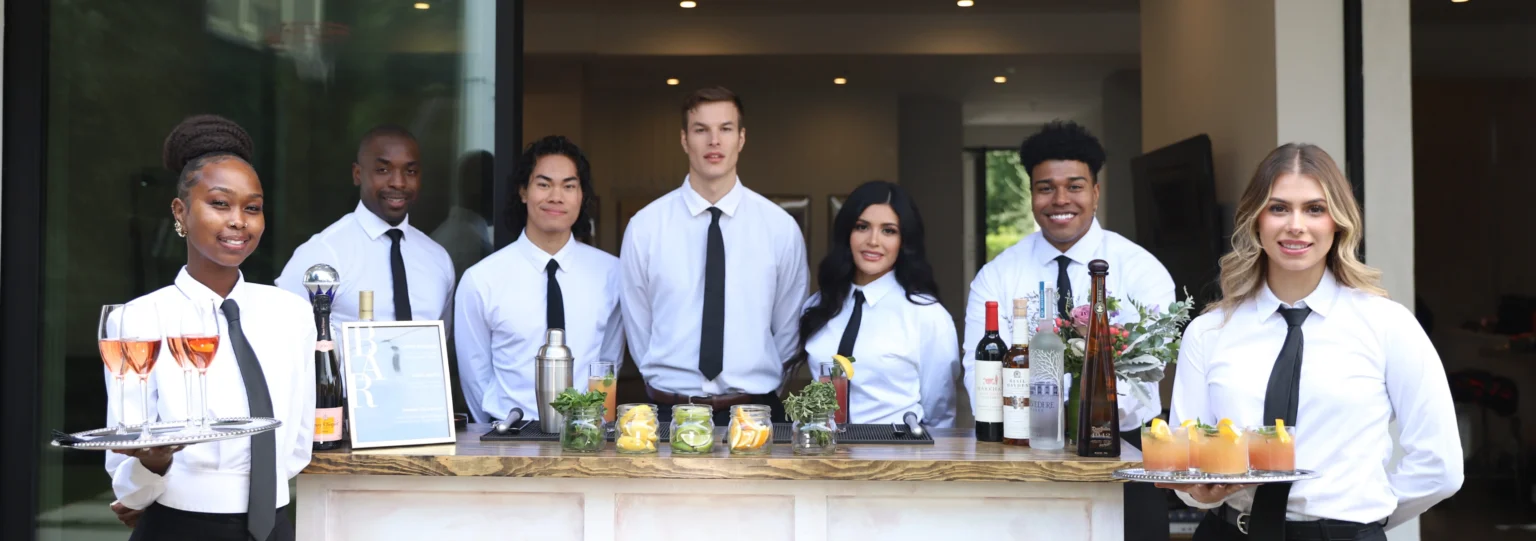 A group of Premier Staff bartenders and servers pose behind a bar counter with various beverages and garnishes.